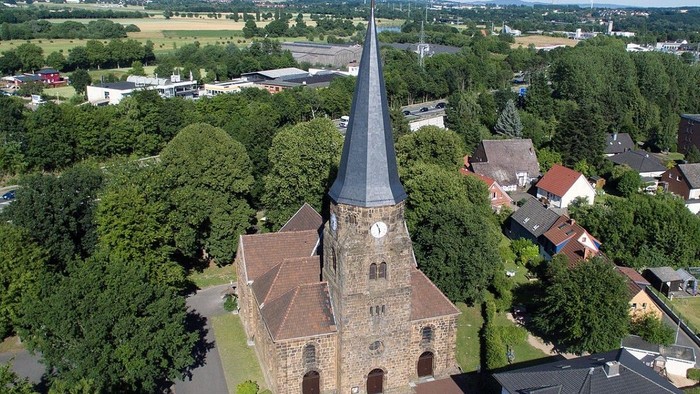 Luftaufnahme einer historischen Steinkirche mit hohem Turm, umgeben von grünen Bäumen und suburbanen Gebäuden.