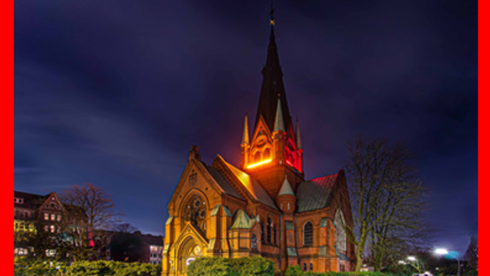 Kirche, die nachts mit rotem Licht beleuchtet wird, umgeben von Bäumen und Gebäuden.