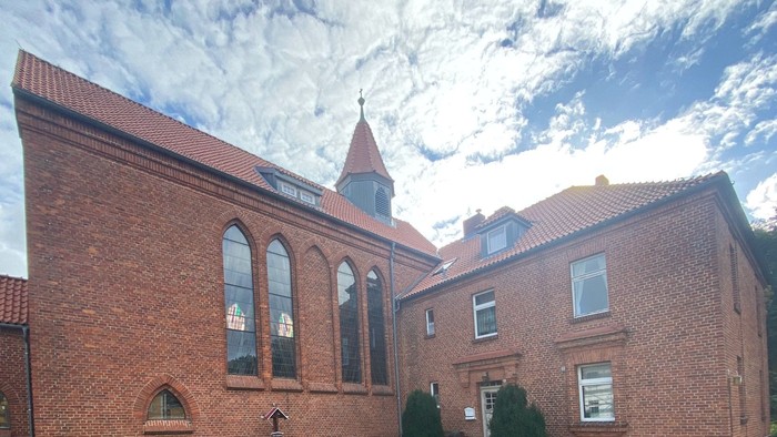 Ein rotes Backsteingebäude mit spitzem Turm und bogenförmigen Fenstern unter einem bewölkten Himmel.
