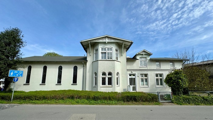 Ein großes, weißes Haus mit zahlreichen Fenstern und einem Tor im Vorgarten.