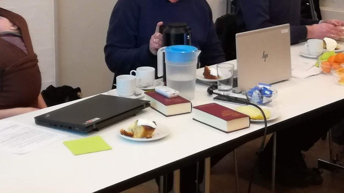 To dagslys og en gruppe af mennesker sidder ved bord med laptopter, noter og kaffe.