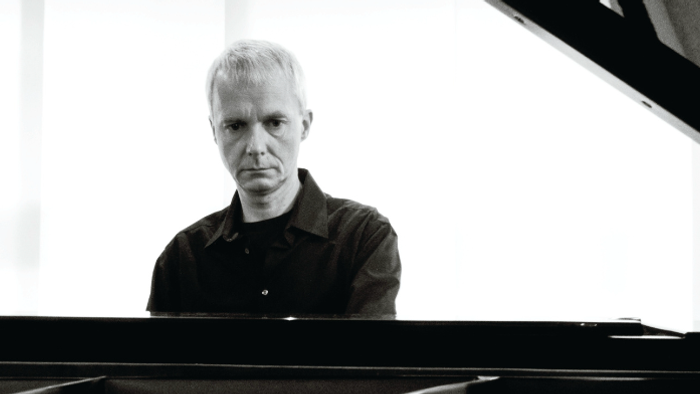 Ein Mann mit kurzem weißen Haar spielt konzentriert Klavier in einem minimalistischen Studio.