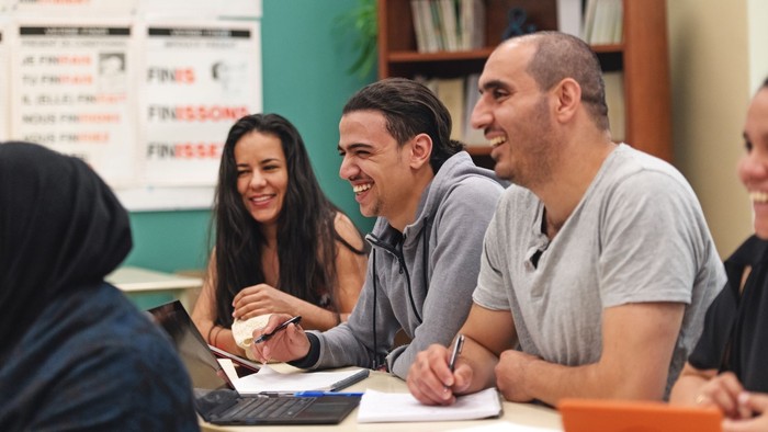 Ein Gruppe von Menschen lacht und arbeitet an einem Tisch in einem Klassenzimmer.