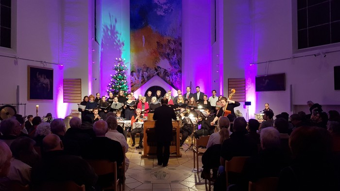Ein Orchester spielt in einem großen Saal vor einem Weihnachtsbaum und einem Publikum.