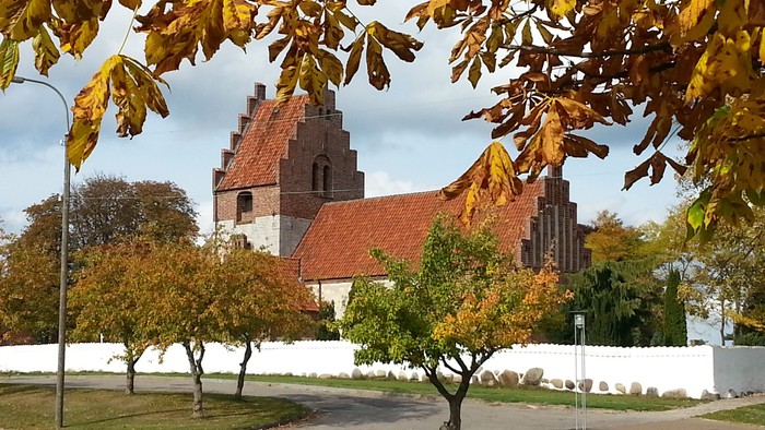 En kirke med røde tag og en mur, omgivet af faldne blade og træer