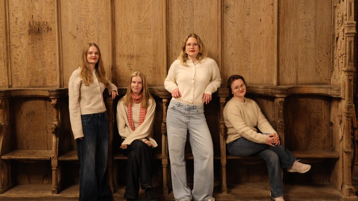 Fire kvinder poser sammen, tre sidder og en står, foran en trævægsbaggrund.