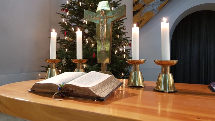 Ein hölzerner Tisch mit offener Bibel, vier brennenden Kerzen und einem Weihnachtsbaum im Hintergrund