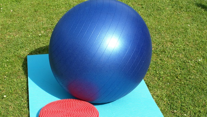 Ein blauer Fitnessball und ein rotes Übungsgerät liegen auf einer blauen Matte im Gras.