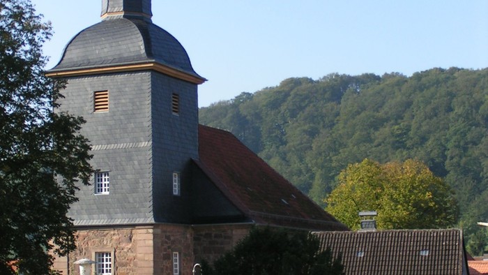 Kirche mit hohem Kirchturm und Kreuz, umgeben von Bäumen und Häusern.