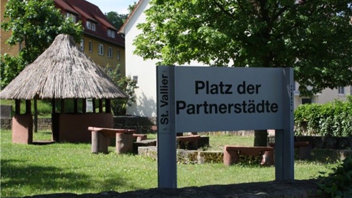 Ein Schild im Park trägt die Aufschrift *„Platz der Partnerstädte“* mit Gebäuden und Bäumen im Hintergrund.