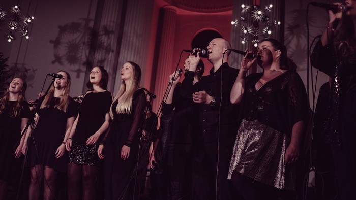 En gruppe sanger på scenen, alle i mørke klæder og med mikrofoner.