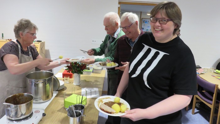 En gruppe mennesker serverer og spiser mad i en køkkenmiljø.