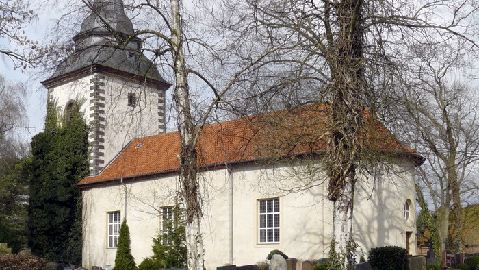 Kleine weiße Kirche mit hohem Turm und rotem Dach in ländlicher Umgebung.