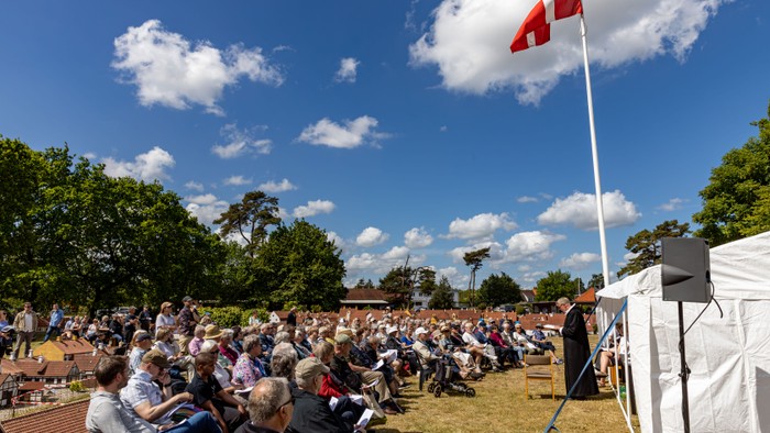 Udendørs samling med en folkemængde siddende på græsset under et flag, der lytter til en taler nær et hvidt telt.