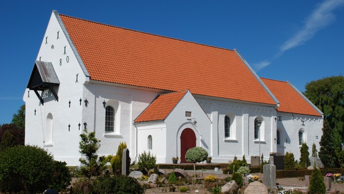 Hvid kirke med rødt tag og rød dør, omgivet af grønne planter og gravestene.