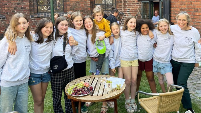 En gruppe unge piger samlet om en bord med mad og drikke i en have foran et rødt murværk.