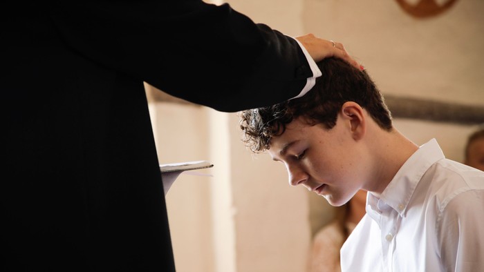 En ung dreng med lukkede øjne og kræmte hår får blessing fra en præst.