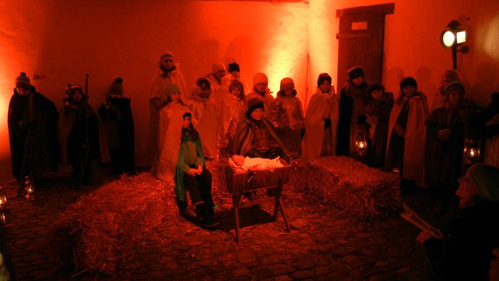 Eine Gruppe von Menschen in Gewändern versammelt sich um eine Krippe, in der ein Baby liegt, bei Kerzenlicht.