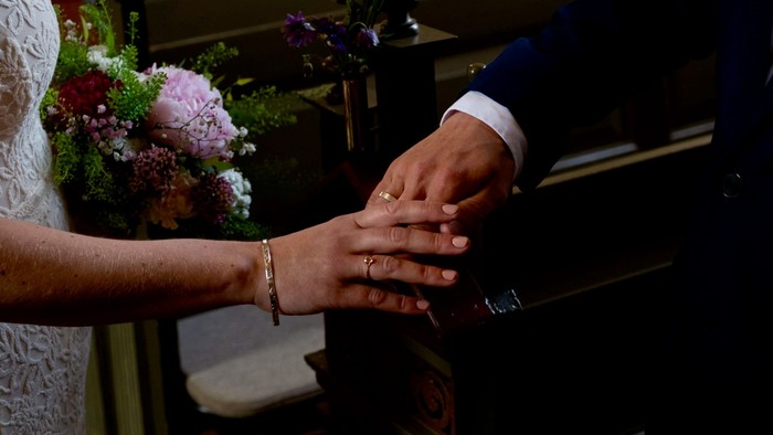 Par, der holder hinanden i hånden under en bryllupsceremoni med blomsterdekorationer i baggrunden.