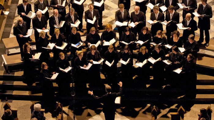 Ein großer Chor tritt in einem prächtigen, schwach beleuchteten Konzertsaal auf, während ein Dirigent von vorne leitet.