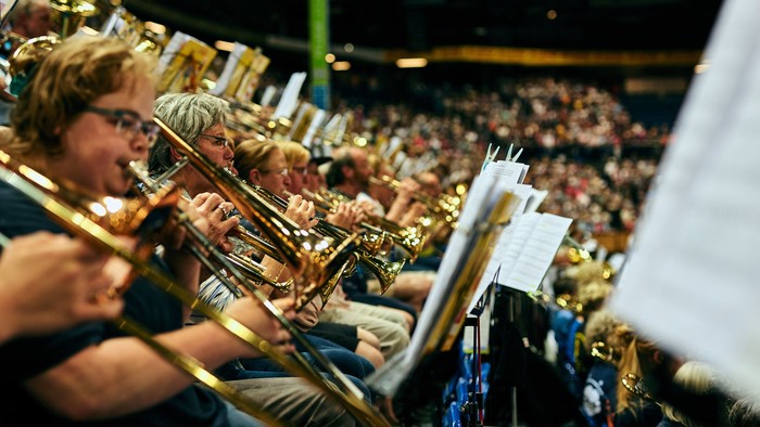 Eine Blaskapelle spielt energiegeladen in einem vollbesetzten Konzertsaal, Trompeten und Posaunen erklingen.