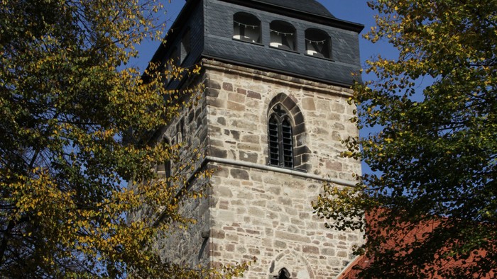 Steinerner Kirchturm mit rotem Dach und Uhr, umgeben von Bäumen.