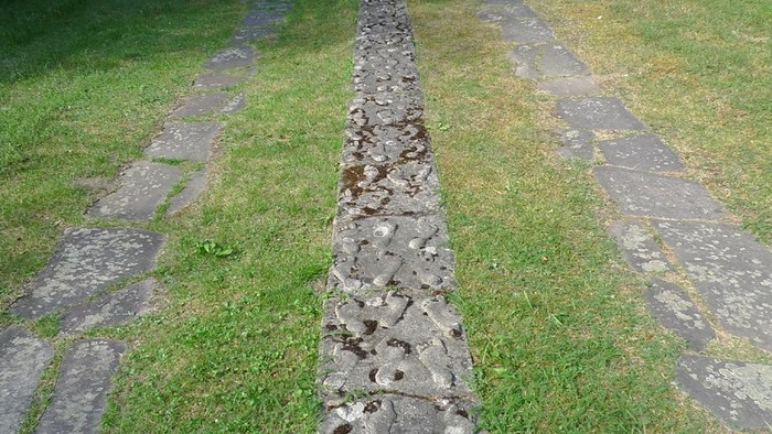 Eine verwitterte Steinmauer verläuft durch einen grasbewachsenen, gepflasterten Weg in einer üppigen Außenanlage.