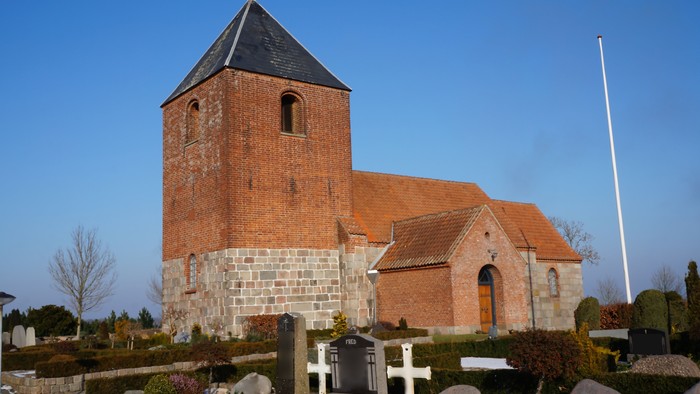 En lille kirke med tårn og røde tag, omgivet af en gravplads og en klar blå himmel