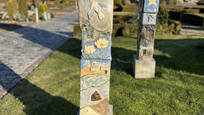 Farverig totempæl i græsbevokset område med bygning og tårn i baggrunden.