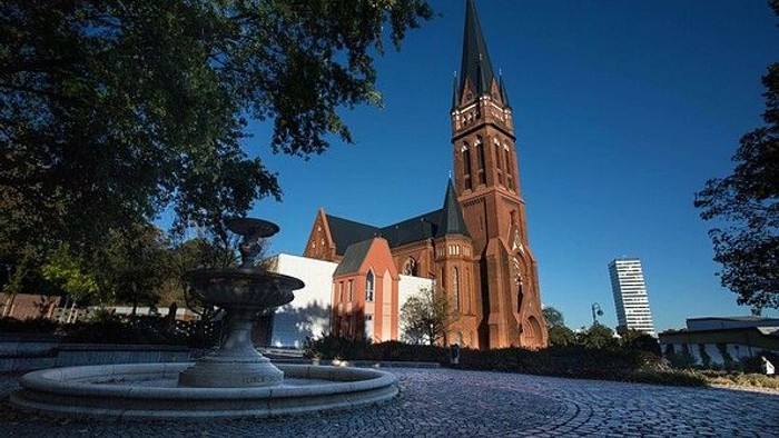 Im Vordergrund ein Brunnen, dahinter eine rote Kirche mit hohem Turm und im Hintergrund ein modernes Gebäude