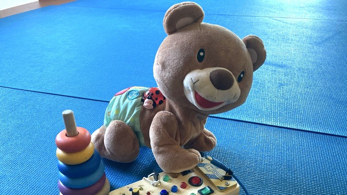 Ein brauner Teddybär sitzt auf einer blauen Matte neben einer Spielzeugtastatur und einem bunten Stapelspielzeug.