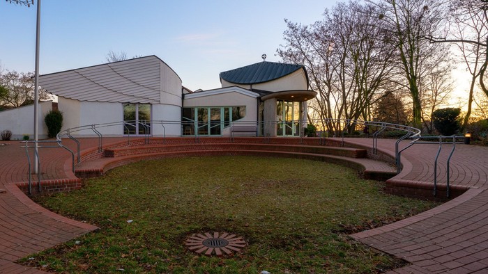 Moderner Gebäudekomplex, umgeben von einem gestalteten Garten mit Ziegelsteinwegen und Grünflächen bei Sonnenuntergang.