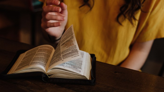 Eine Person in gelbem Kleid liest ein aufgeschlagenes Buch auf einem Holztisch.