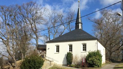 Kleine weiße Kirche mit schwarzem Dach und Turm, umgeben von Bäumen und Büschen.