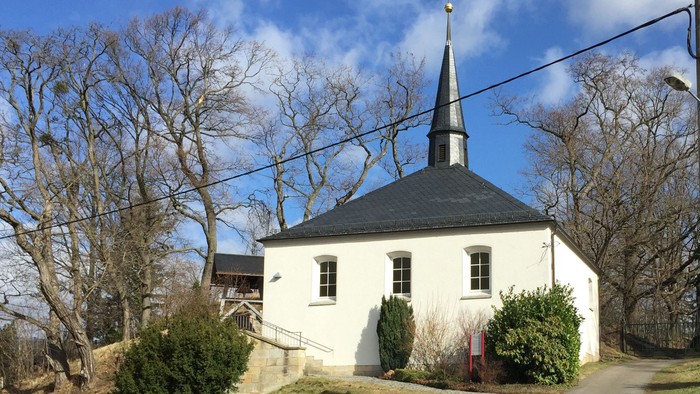 Kleine weiße Kirche mit schwarzem Dach und Turm, umgeben von Bäumen und Büschen.