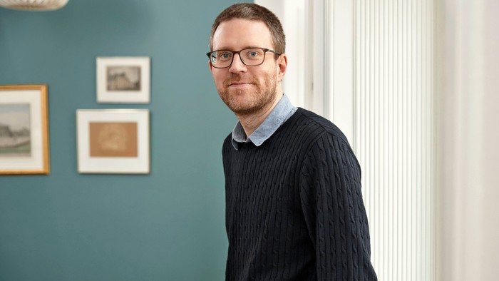 Mand med briller og sweater står nær en væg med indrammede billeder.