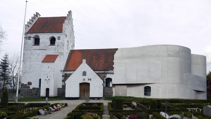 Historisk kirke med moderne arkitektonisk tilbygning omgivet af en omhyggeligt vedligeholdt havekirkegård.