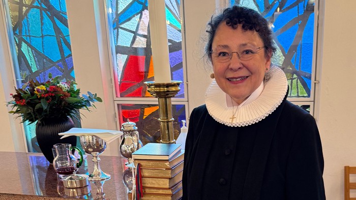 En person i religiøst tøj står ved et kirkealter med farvede glasvinduer bag sig.