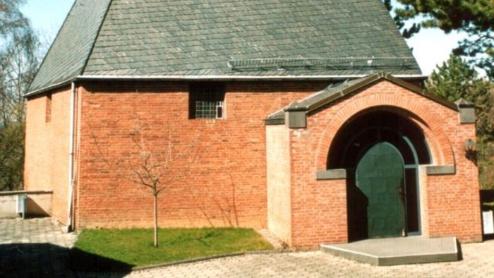 Idyllische Backsteinkapelle mit steilem dreieckigem Dach und markantem Kreuz an der Spitze.
