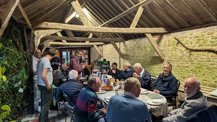 Group of people gathered around tables under wooden structure.