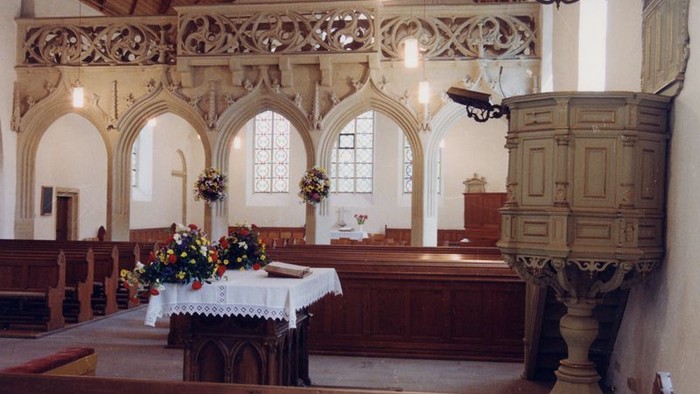 Innenraum einer Kirche mit hölzernen Bänken und einem Altar mit Blumen.