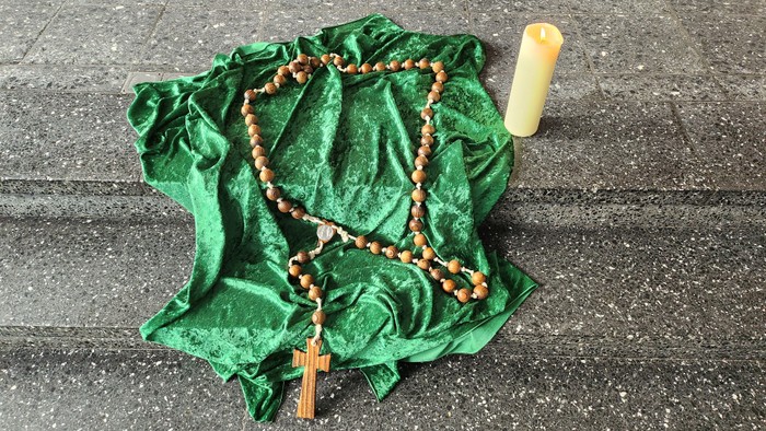 Ein Rosenkranz liegt auf einem grünen Tuch neben einer Kerze auf dem Boden.
