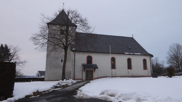 Eine kleine, schneebedeckte Kirche mit einem hohen Glockenturm steht in einer ruhigen Winterlandschaft.
