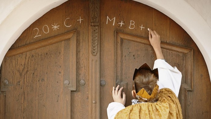 Ein Priester in goldener Robe klopft an eine hölzerne Tür mit eingravierten Buchstaben und Zahlen.