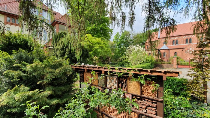 Ein hölzerner Steg führt durch einen üppigen Garten mit hohem Grün und blühenden Pflanzen, umgeben von roten Gebäuden.
