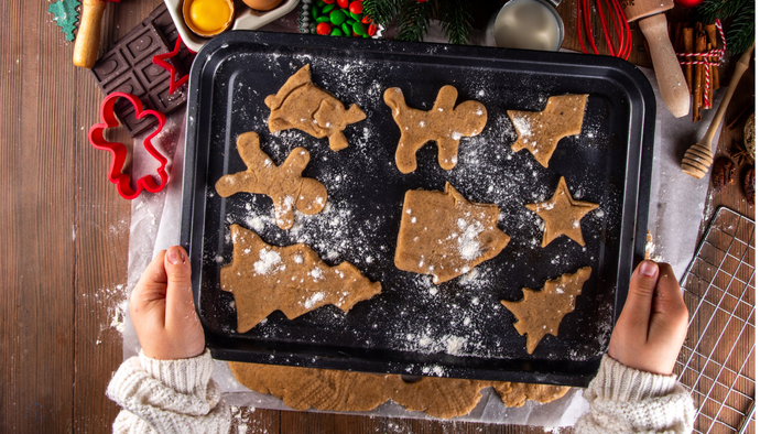 Hände halten ein Tablett mit Lebkuchen, umgeben von Backzutaten und Weihnachtsdekoration.