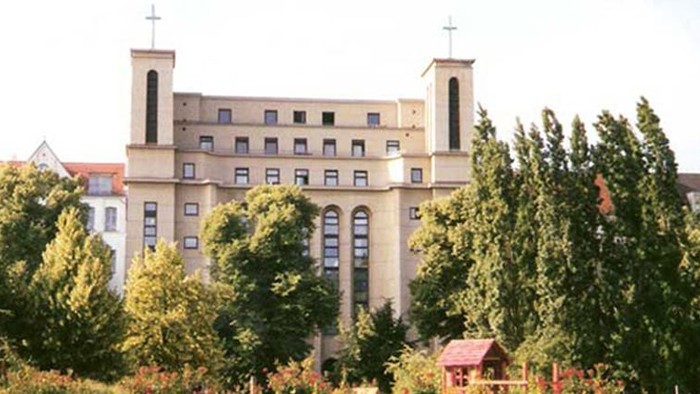 Großes, mehrstöckiges Gebäude mit zwei Türmen, umgeben von Bäumen und Büschen