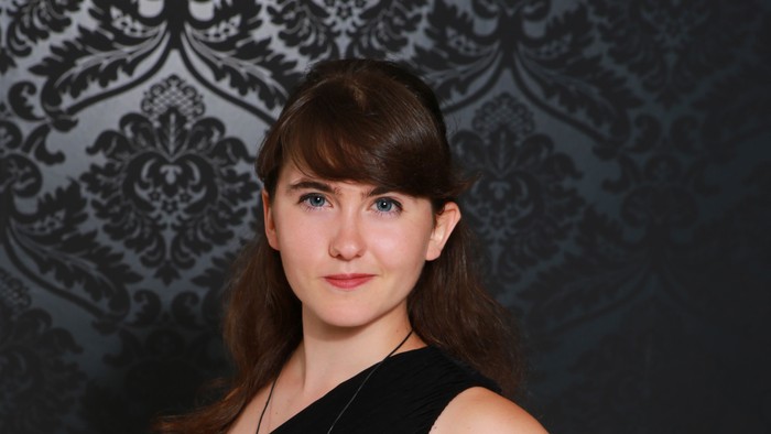 Eine Frau in einem ärmellosen schwarzen Kleid posiert selbstbewusst vor einem verzierten dunklen Hintergrund.