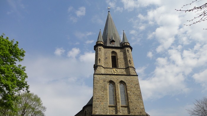 Ein hoher Kirchturm ragt vor einem klaren blauen Himmel auf, flankiert von Bäumen.