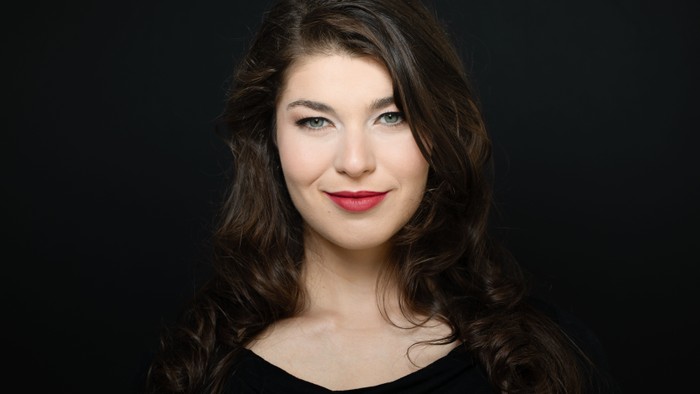 Porträt einer Frau mit langen dunklen Haaren, die ein schwarzes Oberteil trägt, vor dunklem Hintergrund.